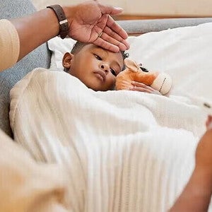 Ένα παιδί ξαπλωμένο με κουβέρτα και θερμόμετρο, φροντίζεται από ενήλικα.