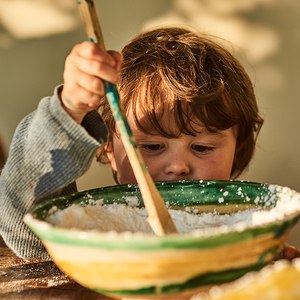 Photograph of a child stirring green paint in a bowl, warm sunlight.