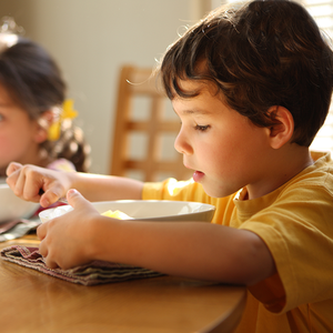 Photograph of two children at breakfast—boy in yellow eating cereal, girl with bow blurred.