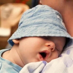 Μωρό με γαλάζιο καπέλο κοιμάται στην αγκαλιά ενήλικα.