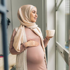 Photograph of woman wearing a hijab, cradling her abdomen and holding a mug, gazing out window