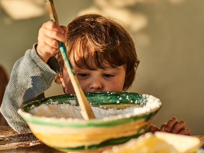 Photograph of a child stirring green paint in a bowl, warm sunlight.