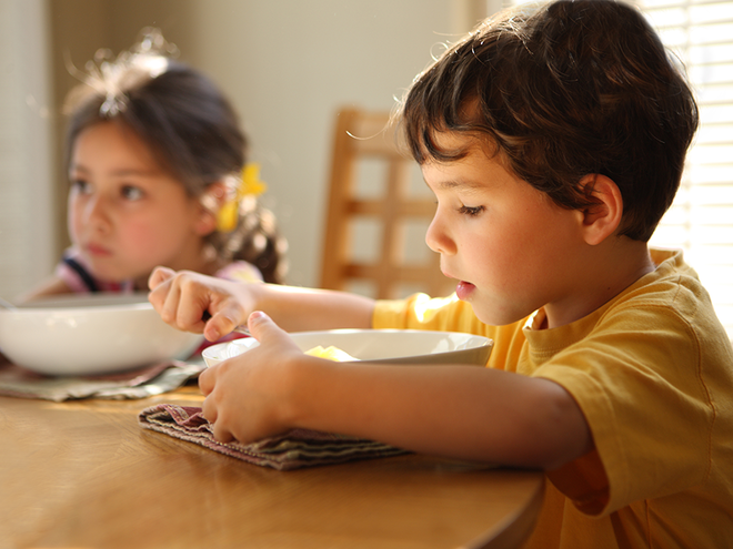 Photograph of two children at breakfast—boy in yellow eating cereal, girl with bow blurred.