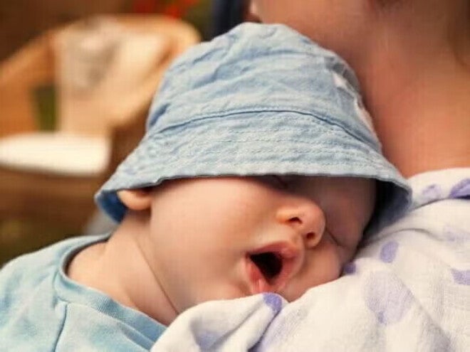 Μωρό με γαλάζιο καπέλο κοιμάται στην αγκαλιά ενήλικα.