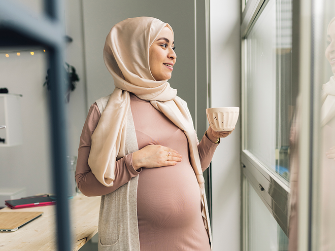 Photograph of woman wearing a hijab, cradling her abdomen and holding a mug, gazing out window