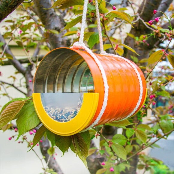 Μία πορτοκαλί κονσέρβα στο ρόλο ταΐστρας πουλιών κρέμεται με σχοινί από ένα δέντρο. Μία πορτοκαλί κονσέρβα στο ρόλο ταΐστρας πουλιών κρέμεται με σχοινί από ένα δέντρο.